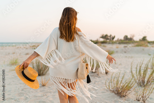 Summer mood. Playful brunette woman dancing in sunlight on the beach. Boho style. Trendy necklase and earrings.  View from back. photo