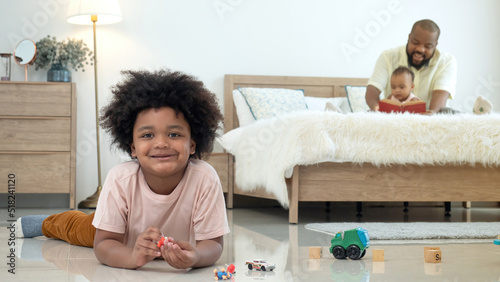 Little African boy lying down playing toys on the floor, smile and look at camera with blurry background of father reading book for little sibbling sister on the bed. photo