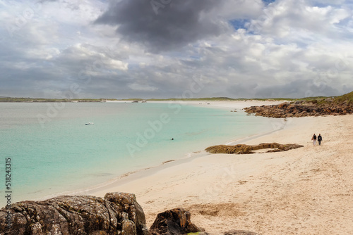 Couple walking on a sandy beach with rocks by a beautiful turquoise color ocean water. Gurteen beach  county Galway  Ireland. Popular tourism area with amazing nature scene. Irish landscape.