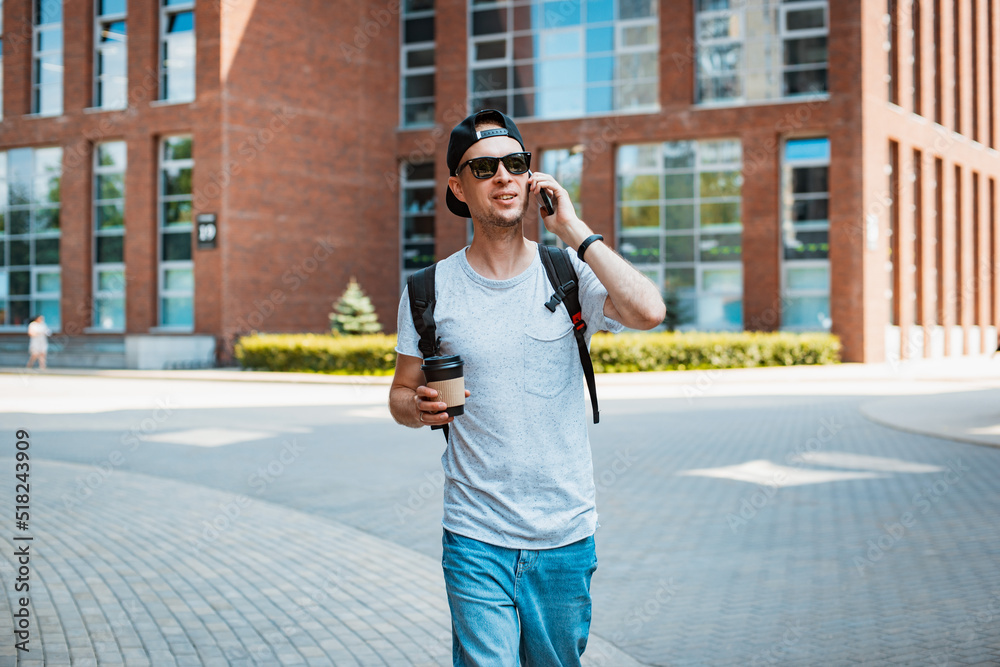 Fototapeta premium young man is a fashionable stylish guy in sunglasses and a black cap walking around the city holding a smartphone in his hand and listening to music headphones. T