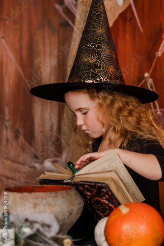 Cute red-haired girl in a black witch costume and hat is reading a book in Halloween decorations.Halloween concept.Selective focus.
