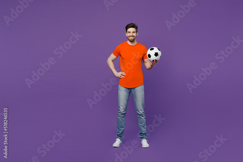 Full body young smiling cheerful fan man he wear orange t-shirt cheer up support football sport team hold in hand soccer ball watch tv live stream look camera isolated on plain dark purple background.