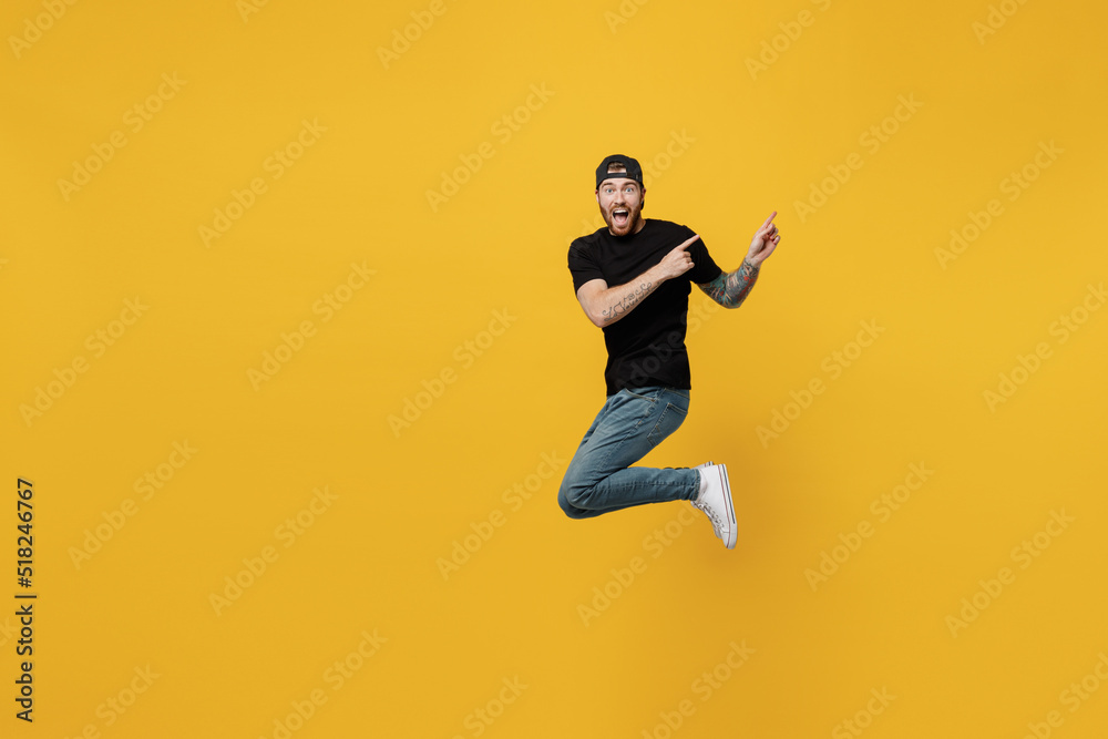 Full body young bearded tattooed man 20s he wears casual black t-shirt cap jump high indicate point index finger aside on workspace area isolated on plain yellow wall background. Tattoo translate fun.