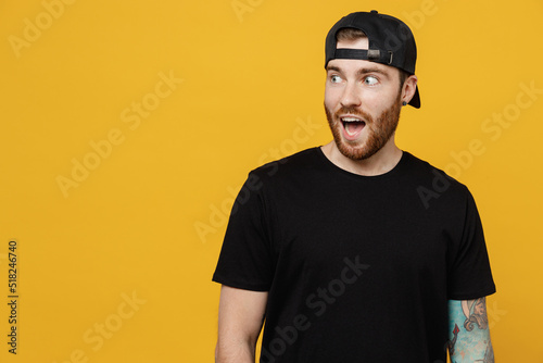 Young surprised fun amazed bearded tattooed man 20s he wears casual black t-shirt cap look aside on workspace area mock up copy space with opened mouth isolated on plain yellow wall background studio