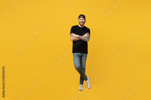 Full body young smiling bearded tattooed man 20s he wears casual black t-shirt cap hold hands crossed folded look camera isolated on plain yellow wall background studio portrait. Tattoo translate fun.