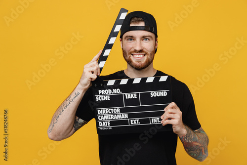 Young smiling bearded tattooed man 20s he wears casual black t-shirt cap holding classic black film making clapperboard isolated on plain yellow wall background studio portrait. Tattoo translate fun.