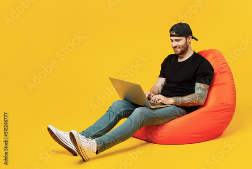 Full body young bearded tattooed man 20s he wear casual black t-shirt cap sit in bag chair hold use work on laptop pc computer isolated on plain yellow wall background studio People lifestyle concept