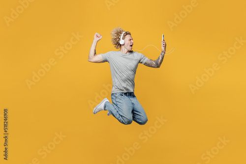 Full body side view young cool excited caucasian man 20s he wear grey t-shirt headphones listen to music do winner gesture jump high isolated on plain yellow backround studio People lifestyle concept © ViDi Studio