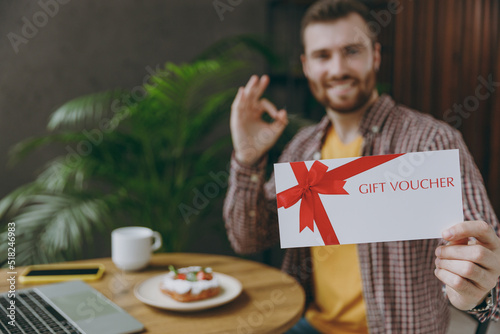 Young happy man he wear shirt hold gift store certificate coupon voucher card show ok sit alone at table in coffee shop cafe relax in restaurant in free time. Freelance mobile office business concept.