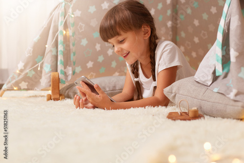 Side view portrait of cute little girl with pigtails wearing white t shirt posing in wigwam at home and using phone, looking at display with smile, enjoying her leisure time at home in peetee tent. photo