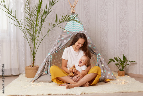 Image of happy laughing baby girl and her mother playing at home in wigwam while sitting on the floor, kid holding wooden toy, mommy enjoying her time with charming daughter. photo