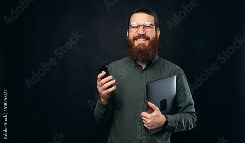 A happy bearded man is holding a laptop and his phone and is smiling is looking at the camera