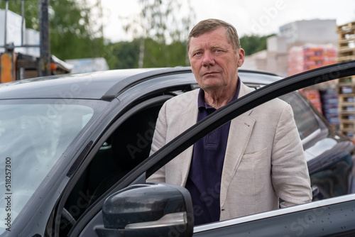 Thoughtful mature man in formalwear looking at camera near car. Serious senior man standing next to car at hardware store with building materials for construction