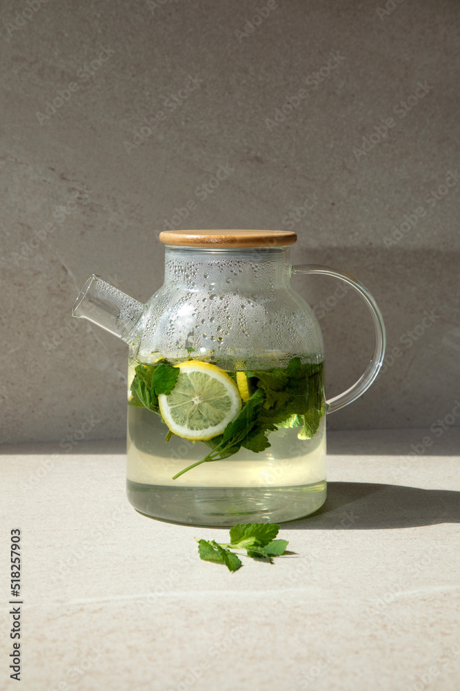Hot tea with mint and lemon in a glass teapot on a light stone table with with shadows. Front view, close up Hot tea with mint and lemon in a glass teapot on a light stone table with with shadows. Front view, close up