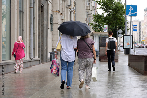 Rain in a city, two women with one umbrella walk on a street on people background. Rainy weather, urban life in summer