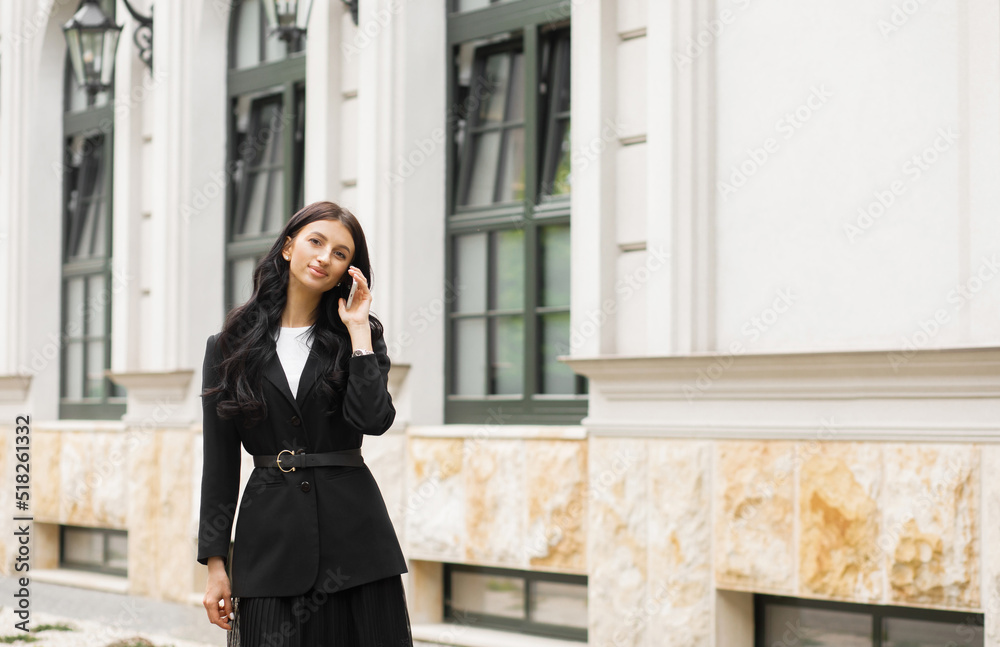 Fototapeta premium Successful businesswoman in a suit with a document goes to a meeting near a modern office building and talks on a smartphone