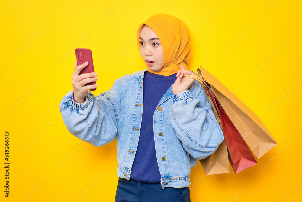 Shocked young Asian woman in jeans jacket using mobile phone for online shopping, holding shopper bags isolated over yellow background