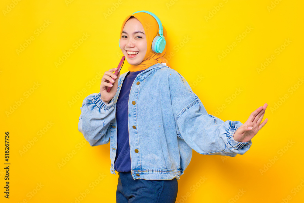 Dancing young Asian woman in jeans jacket wearing headphones for listening to music and singing via mobile phone isolated over yellow background Dancing young Asian woman in jeans jacket wearing headphones for listening to music and singing via mobile phone isolated over yellow background