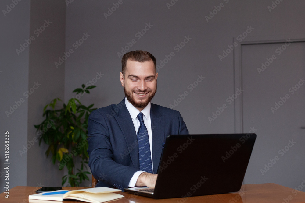 Young happy businessman smiling while using his laptop. Portrait of smiling business man reading message and working in office. Man working at his desk at office. Young happy businessman smiling while using his laptop. Portrait of smiling business man reading message and working in office. Man working at his desk at office.