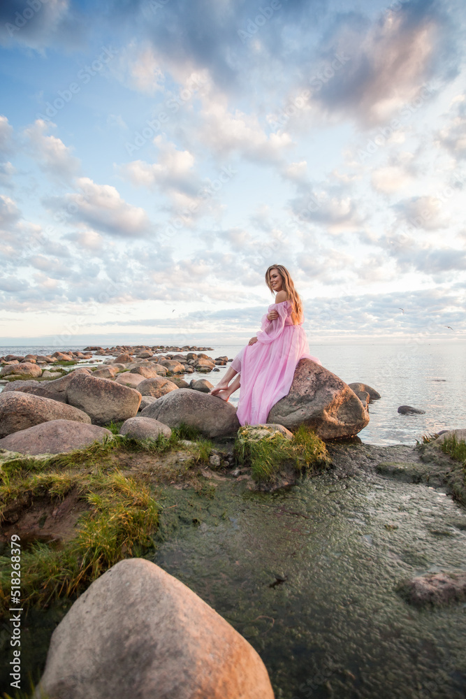 Cheerful glamorous blonde model woman in pink silky summer dress resting against beautiful sea and blue sky with white clouds sunset nature landscape backround