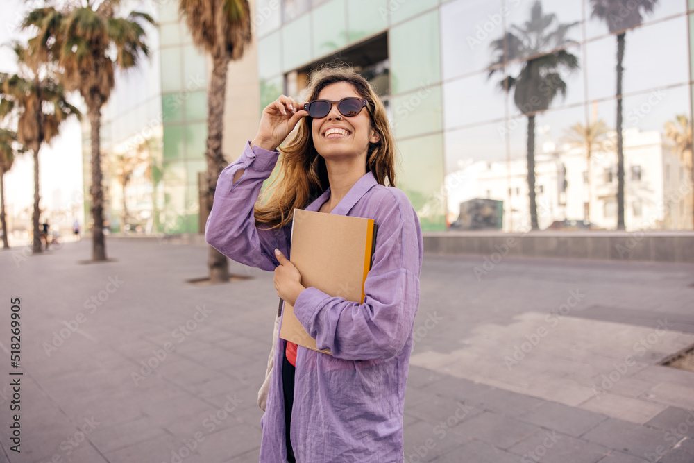 Fototapeta premium Pretty young caucasian woman with notebooks walks around city after class. Brown-haired girl wears sunglasses and purple shirt. Student concept