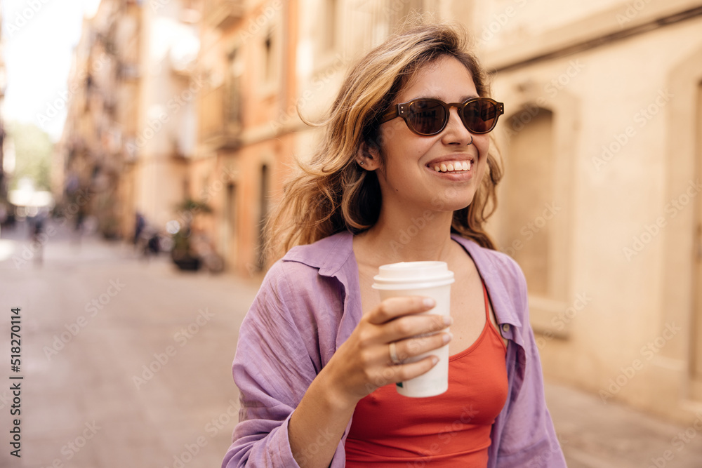 Pretty young caucasian girl with coffee in her hands spends leisure time walking along street morning. Blonde wears sunglasses, casual clothes. City life concept Pretty young caucasian girl with coffee in her hands spends leisure time walking along street morning. Blonde wears sunglasses, casual clothes. City life concept