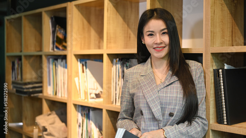 Experienced business woman standing near bookshelves in her personal office and smiling to camera