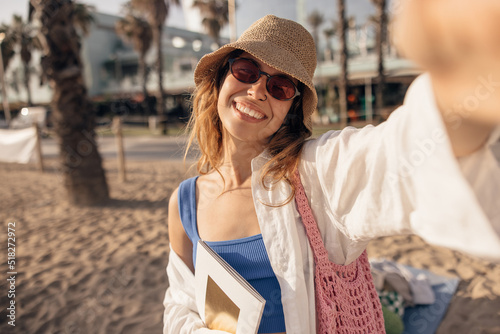 Smiling young caucasian woman taking selfie spending leisure time on beach in sunny weather. Blonde wears hat, sunglasses and shirt. Summer vacation concept