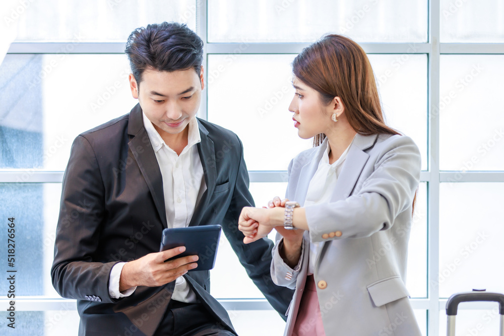Asian young professional successful businessman and businesswoman colleague in formal suit standing together with trolley baggage in airport hallway waiting checking for business trip boarding time Asian young professional successful businessman and businesswoman colleague in formal suit standing together with trolley baggage in airport hallway waiting checking for business trip boarding time
