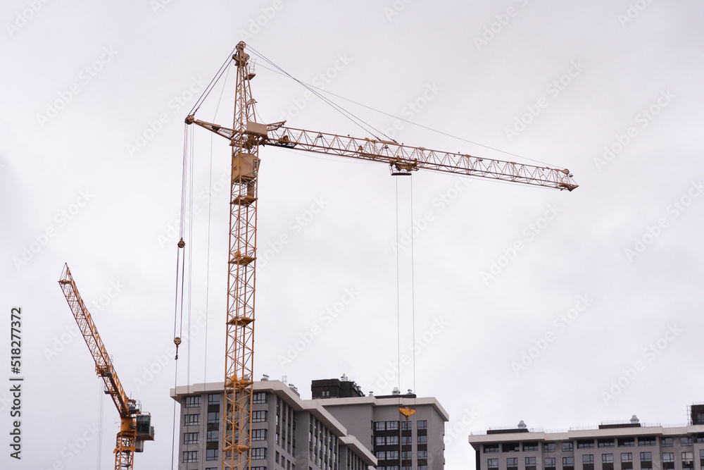 A building site with an orange steel crane on the middle of the frame against cloudy gray sky. A few high houses on the bottom