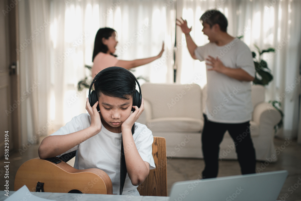Asian boy kid wearing headphones and play loud music. so as not to hear quarrel while parents having fight or quarrel conflict at home. Unhappy problem in family, Domestic problems in the family. Asian boy kid wearing headphones and play loud music. so as not to hear quarrel while parents having fight or quarrel conflict at home. Unhappy problem in family, Domestic problems in the family.