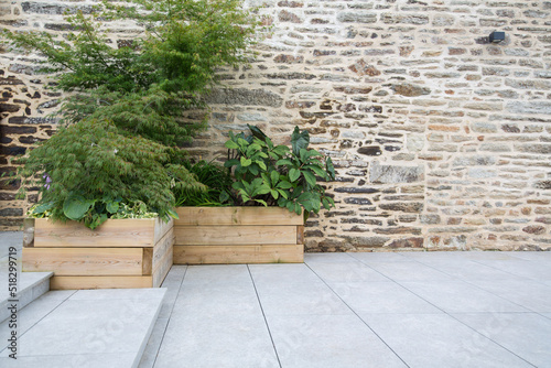 Modern garden design and terrace construction: Hillside plot paved with natural sidewalk flagstones in contrast to raised beds planted with green plants in wooden pots in front of an old stone wall