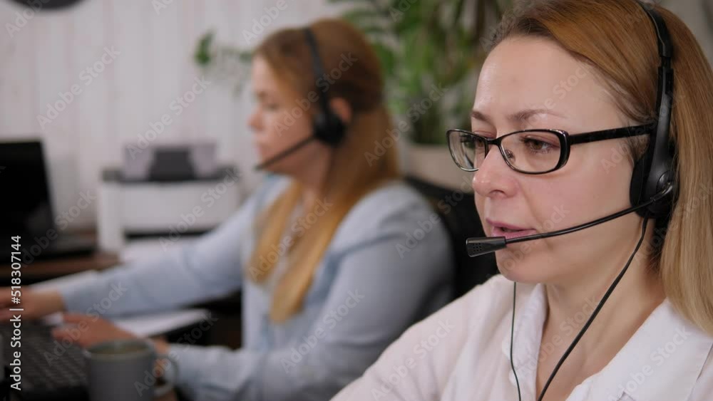 Young positive female customer support phone operator in a microphone headset and glasses working at a computer in the office. Young woman working in a call center surrounded by colleagues. Young positive female customer support phone operator in a microphone headset and glasses working at a computer in the office. Young woman working in a call center surrounded by colleagues.