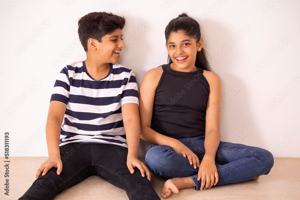 Cheerful boy and girl sitting together on floor against white wall Cheerful boy and girl sitting together on floor against white wall