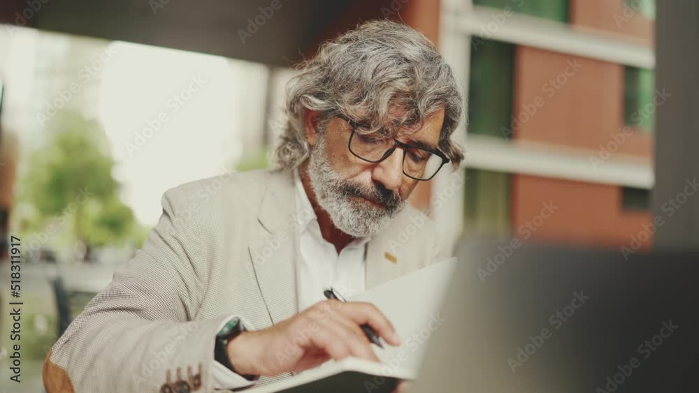 Mature businessman with beard in eyeglasses, user on laptop computer and taking notes, sitting in outdoor cafe. Successful man sitting at table in cafe in nature Mobile office freelancer Mature businessman with beard in eyeglasses, user on laptop computer and taking notes, sitting in outdoor cafe. Successful man sitting at table in cafe in nature Mobile office freelancer