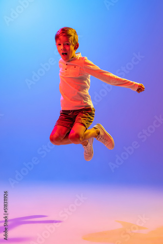 Studio shot of stylish little boy  kid jumping isolated on gradient blue-purple background in neon light. Concept of child emotions  facial expression  fashion and ad.