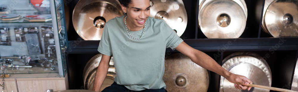 Naklejka premium Cheerful african american customer holding drumstick near plates in music store, banner.