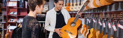 Positive african american seller holding acoustic guitar near woman in music tore, banner.