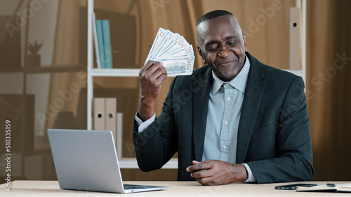 Old mature African businessman 60s worker banker banking agent senior man sitting in office rejoices in financial win holds fan of money sniffing dollars satisfied with salary loan wealth success