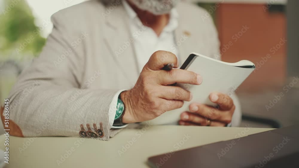Mature businessman with beard in eyeglasses, user on laptop computer and taking notes, sitting in outdoor cafe. Successful man sitting at table in cafe in nature Mobile office freelancer Mature businessman with beard in eyeglasses, user on laptop computer and taking notes, sitting in outdoor cafe. Successful man sitting at table in cafe in nature Mobile office freelancer