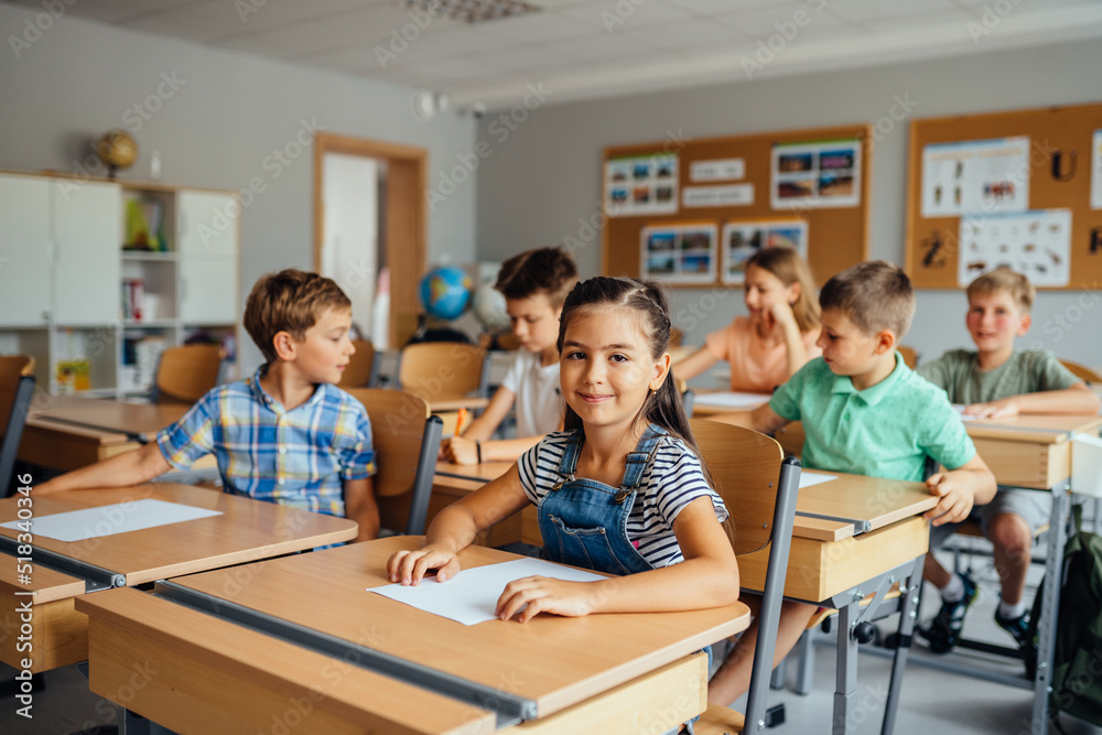 Children in the classroom at the lesson. Young cute hispanic girl looking at the camera. Classmates doing test tasks at school. - obrazy, fototapety, plakaty 