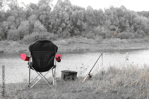 Black and white retro style photo of red fishing chair near fishing rod and fishing bait on river bank in summer on fishing trip