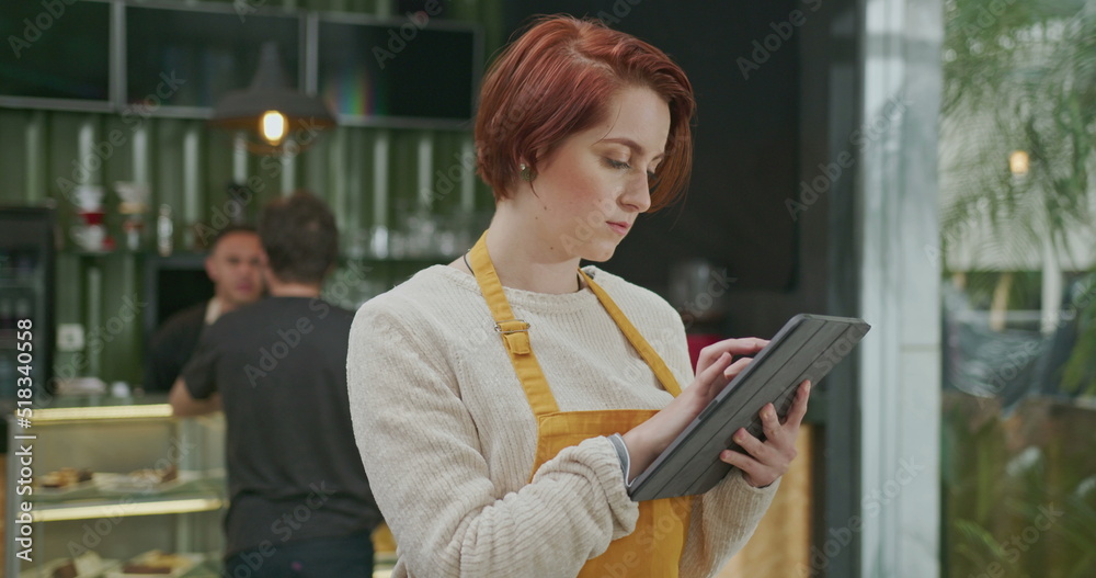 Young employee woman wearing apron standing inside coffee shop holding tablet device checking online orders. Red hair girl working at cafe
