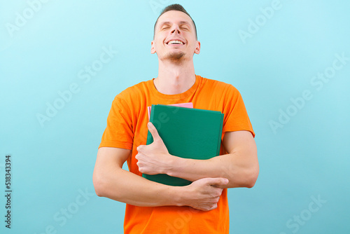 A man in an orange t-shirt holds his head up and smiles, while he is holding files isolated on a white background. Smart. Looking. Office. Business. Happy. Paper. Portrait. Young. Adult. Casual. Cheer © AndriiKoval