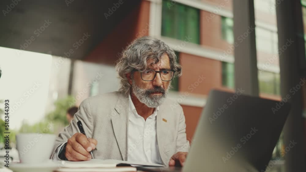 Mature businessman with beard in eyeglasses, user on laptop computer and taking notes, sitting in outdoor cafe. Successful man sitting at table in cafe in nature Mobile office freelancer Mature businessman with beard in eyeglasses, user on laptop computer and taking notes, sitting in outdoor cafe. Successful man sitting at table in cafe in nature Mobile office freelancer