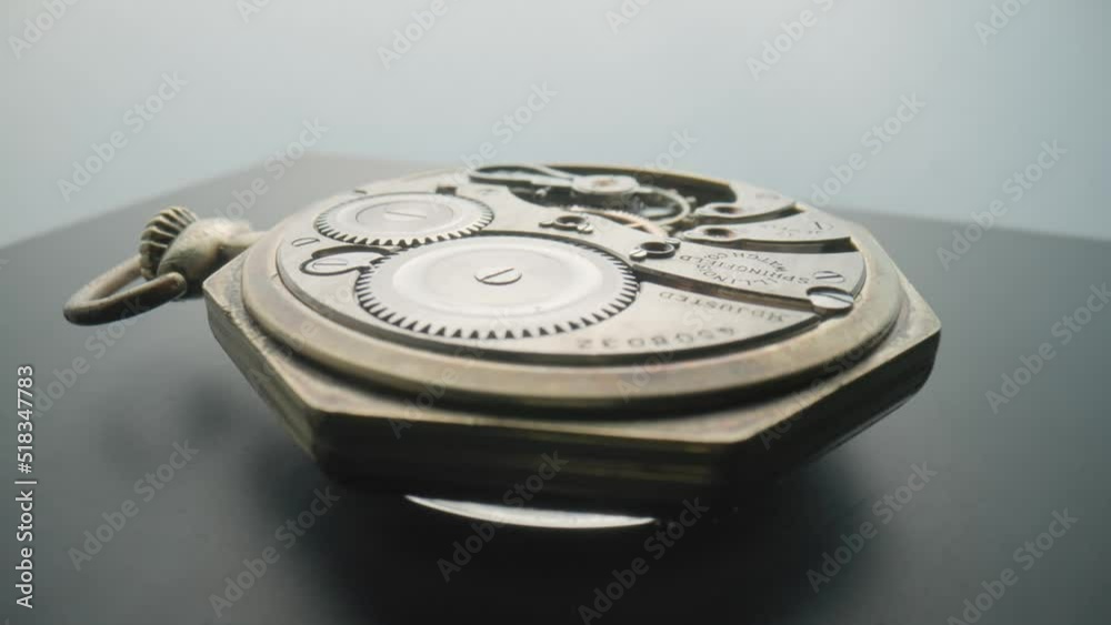 An antique pocket watch with an open working clock mechanism rotates on a gray table in a studio. Reverse side of the clock with rotating gears, spring and gearwheel. Disassembled retro pocket watch.