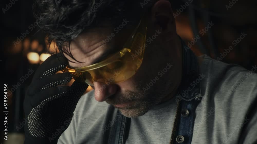 A middle-aged man taking off protective goggles and wiping sweat off his forehead, while doing hard metal grinding work in a garage for his small business.