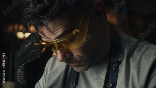 A middle-aged man taking off protective goggles and wiping sweat off his forehead, while doing hard metal grinding work in a garage for his small business.