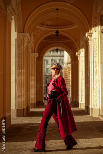 Fashionable confident blonde woman wearing trendy sunglasses, beret, pink turtleneck sweater, dark red autumn coat, flared trousers, walking in street of European city. Outdoor full-length portrait. 
