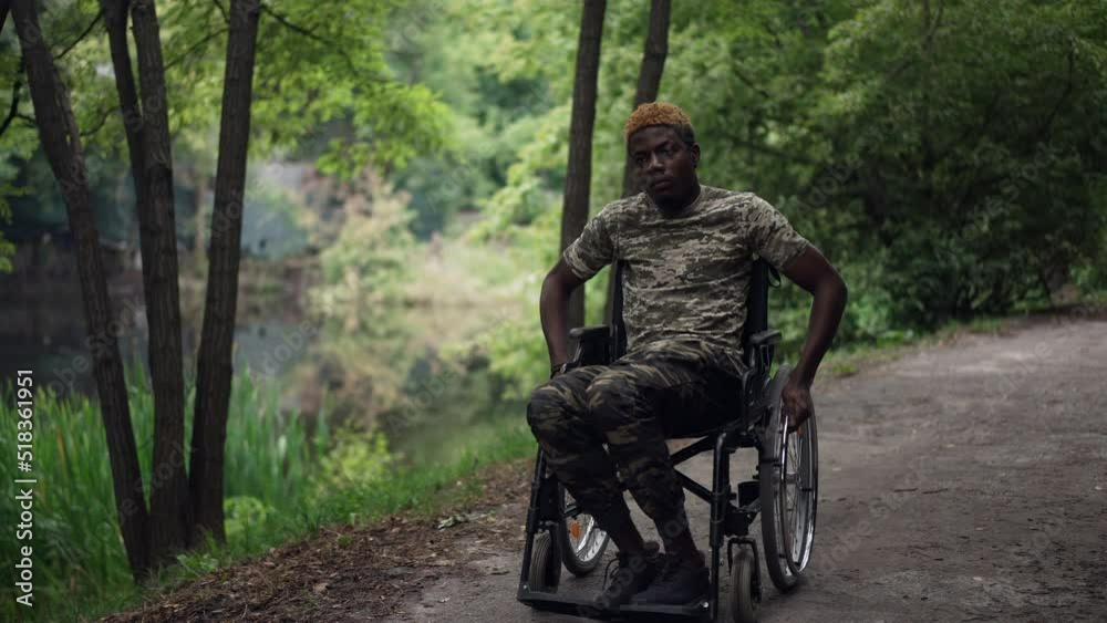 Wide shot sad young soldier in wheelchair riding in summer forest looking away. Front view portrait of African American ill man rolling in park thinking. Depression and procrastination concept Wide shot sad young soldier in wheelchair riding in summer forest looking away. Front view portrait of African American ill man rolling in park thinking. Depression and procrastination concept